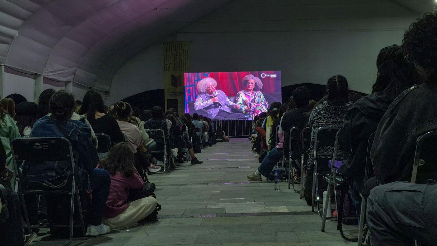 Público sentado frente a una pantalla gigante bajo una carpa.