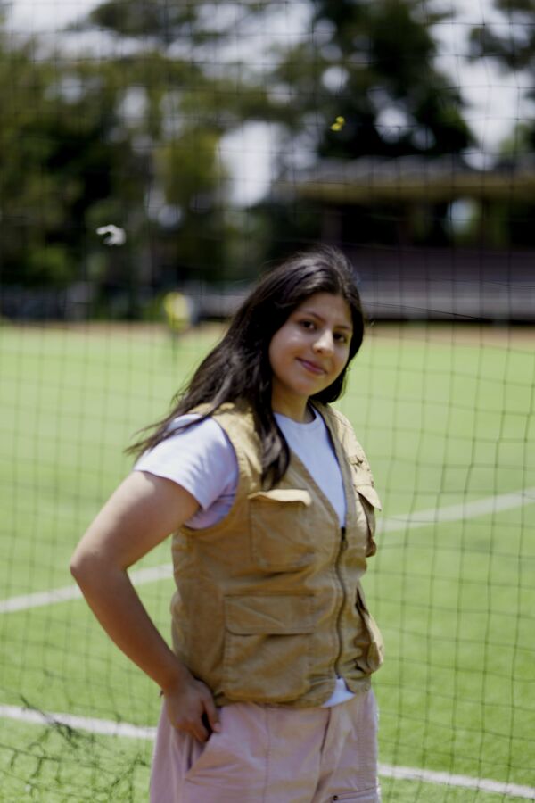 Dania Esparza en una cancha de futbol, con chaleco beige y playera blanca.